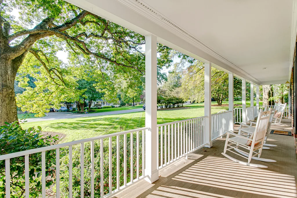 A covered front porch painted white, with a rocking chair, surrounded by large trees.