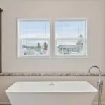 modern, rectangular bathtub with standalone faucet positioned below a wall with dual windows and sitting area