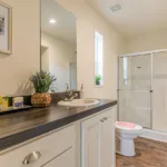 single sink vanity with dark grey counters, nickel hardware, and white cabinets next to a toilet and standing shower with sliding glass doors