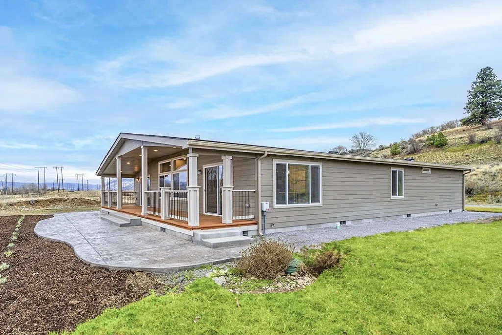 A light gray manufactured home with land, installed on an Oregon property, with a green lawn, front porch, and concrete patio against a blue sky